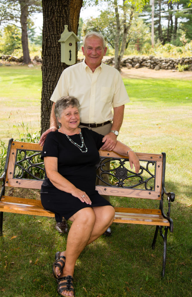 husband standing behind wife, who is sitting on a bench