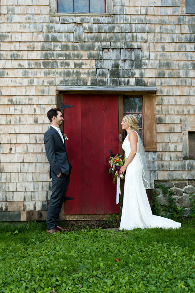 bride and groom standing in front of red barn door looking at each other smiling