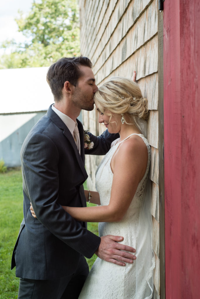 groom kissing bride's head, bride leaning against the barn