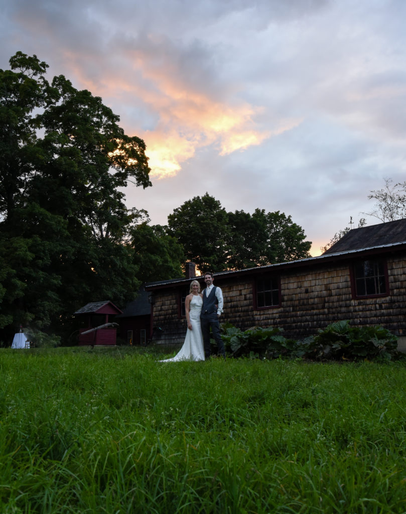 bride and groom standing in front of barn with sunset in background