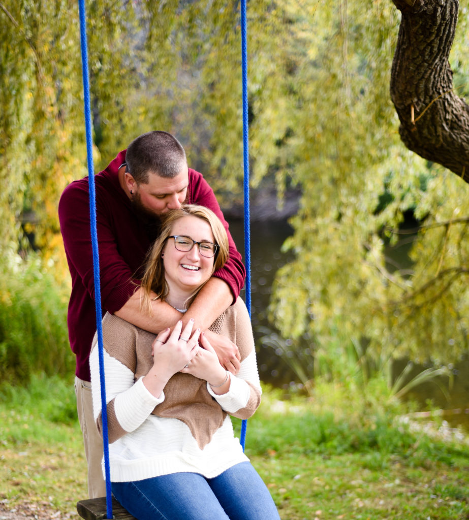 woman sitting on a swing smiling while man hugs her from behind and kisses her on the head