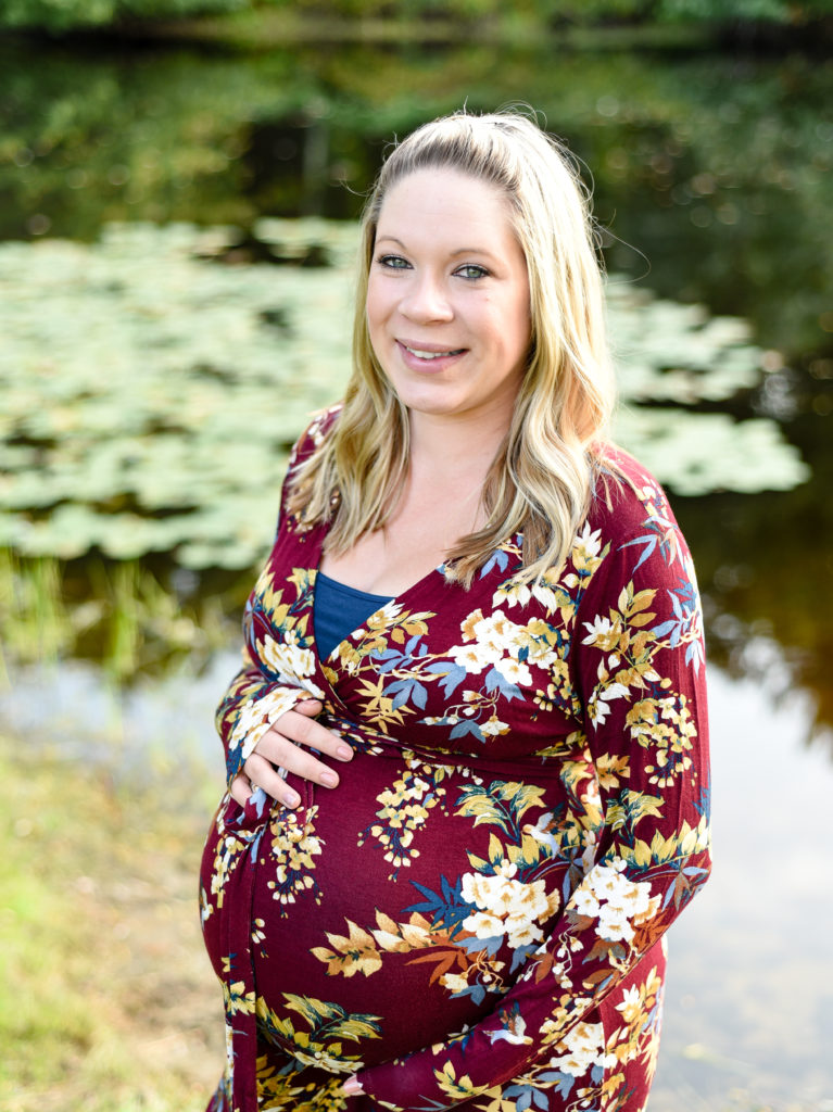 mom to be standing in front of pond holding belly 