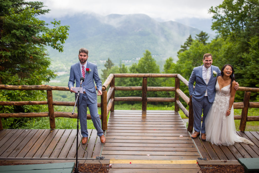 groomsmen giving a speech on deck of mountain top wedding; bride and groom arm in arm listening