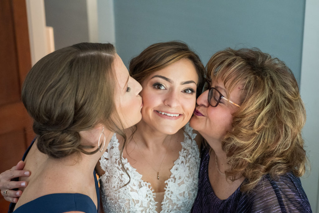 Maid of honor and mother of bride kissing the bride on her cheeks