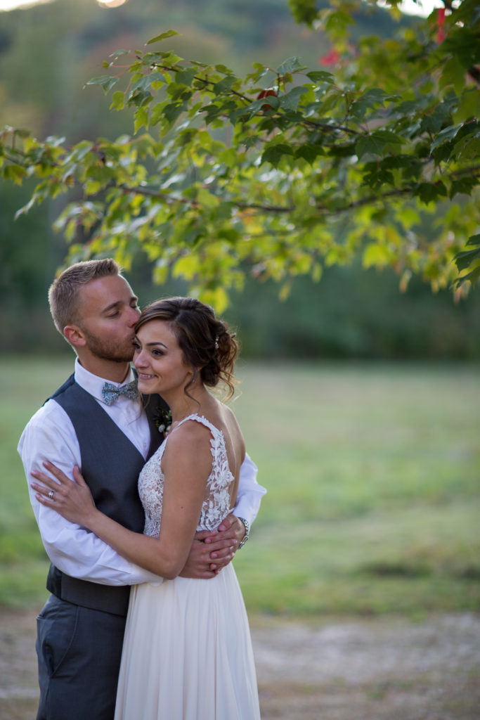 groom kissing bride's forehead while in a hug under a tree at a fall NH wedding