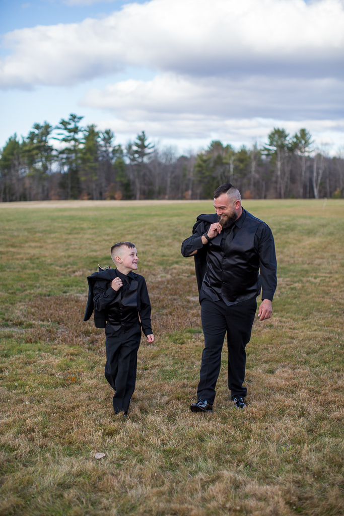 groom and his son walking with their jackets over their shoulders smiling at each other in a field at Whitney Farm Estate