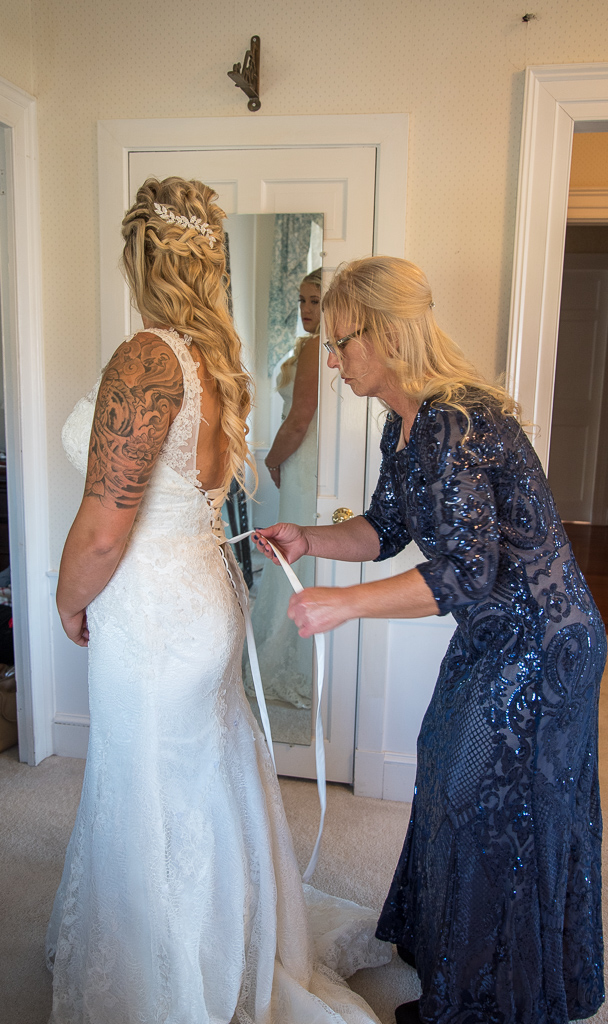 mother of the bride lacing up the back of her dress while bride looks at herself in the mirror