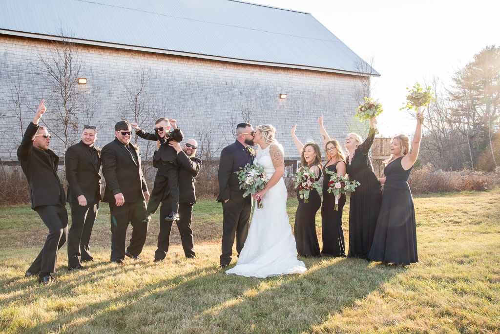 bride and groom kissing with bridal party cheering behind them at Whitney Estate Farm