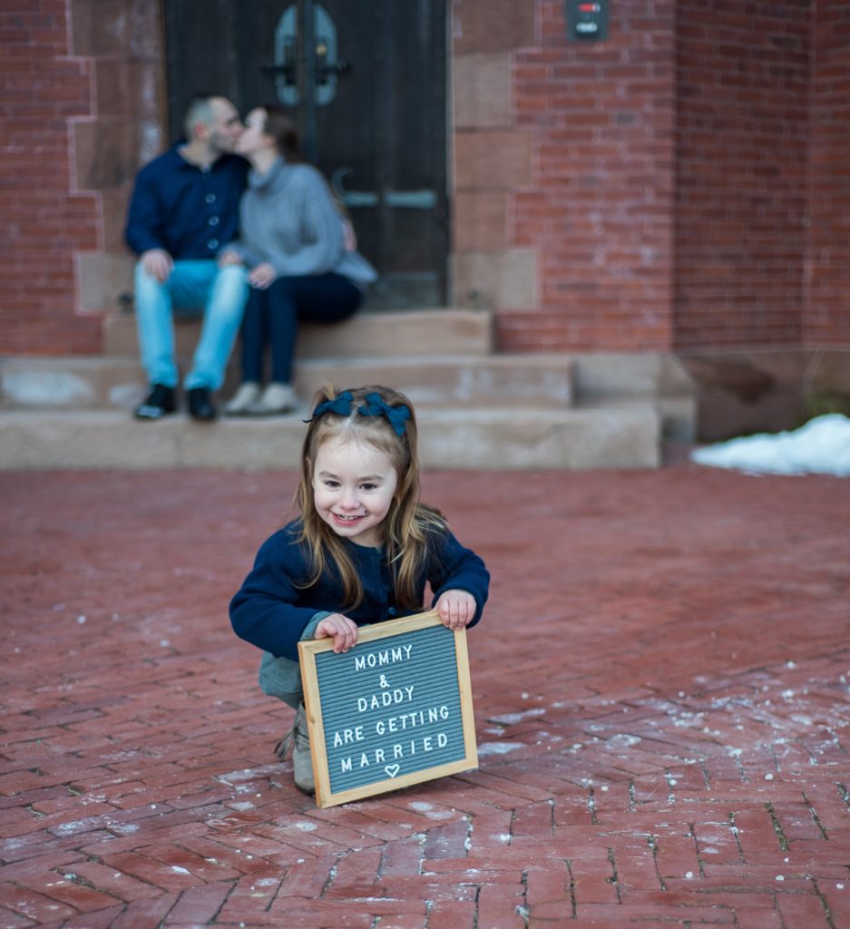 daughter holding a sign "mommy and daddy are getting married" while parents kiss out of focus in the background