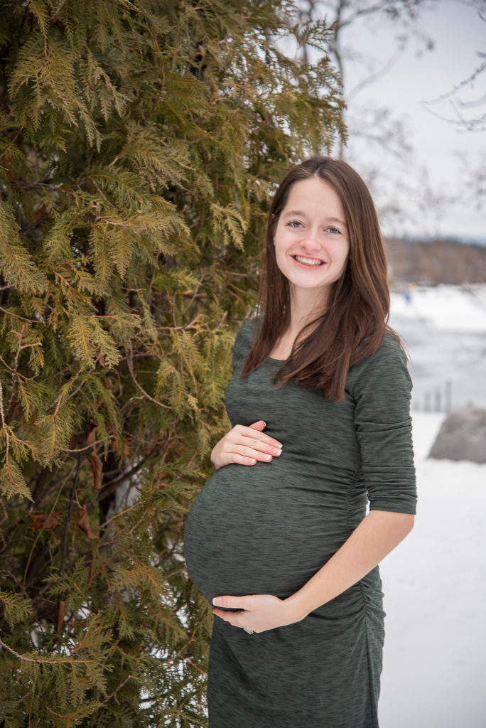 mom to be in front of snowy greenery holding her bump