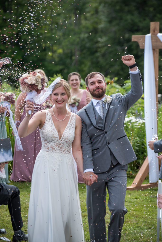 bride and groom walking down the aisle as family throws confetti at them at NH summer barn wedding