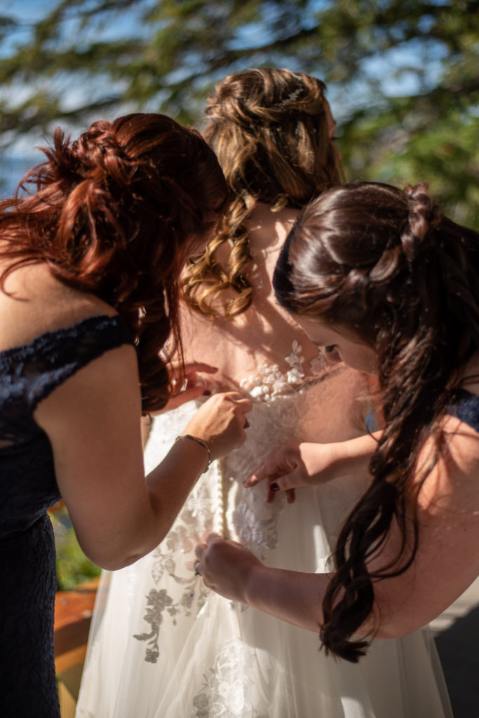 bridesmaids zipping up brides dress at the wedding at the lake