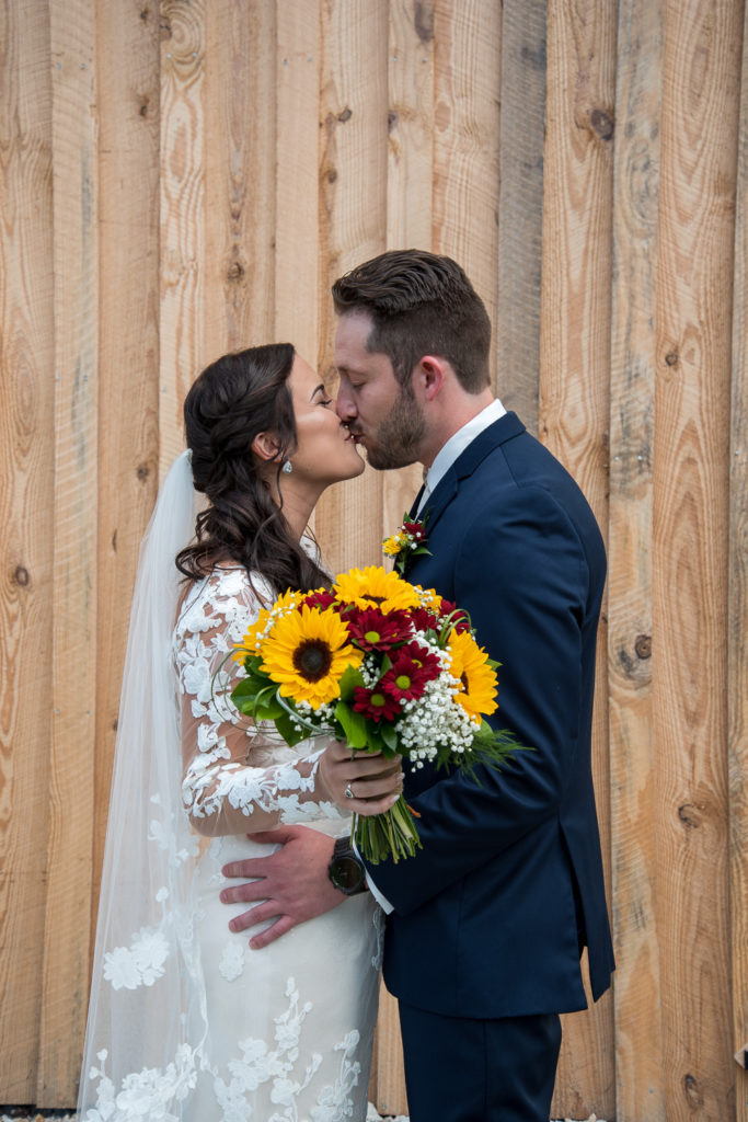 bride and groom kissing in front of barn at wedding rings on a sunflower bouquet at fall wedding at the barn at bull meadow