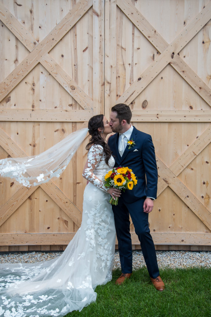 veil flying in the wind while bride and groom kiss at wedding rings on a sunflower bouquet at fall wedding at the barn