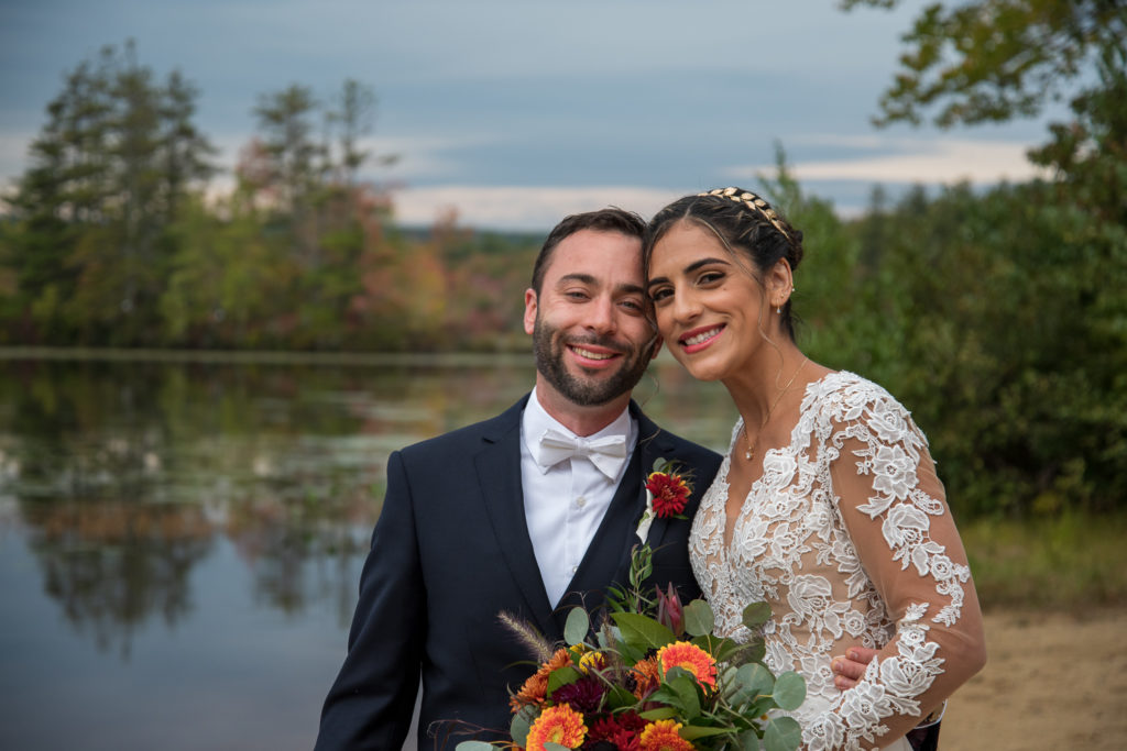 bride and groom posing in front of a pond with fall foliage in the background, shot by NH wedding Photographer