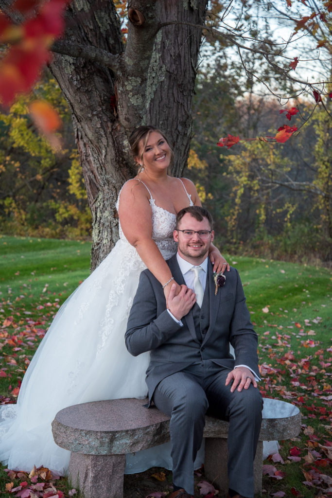 groom sitting on a bench and bride hugging him from behind in front of a red foliage tree at the perfect fall wedding 