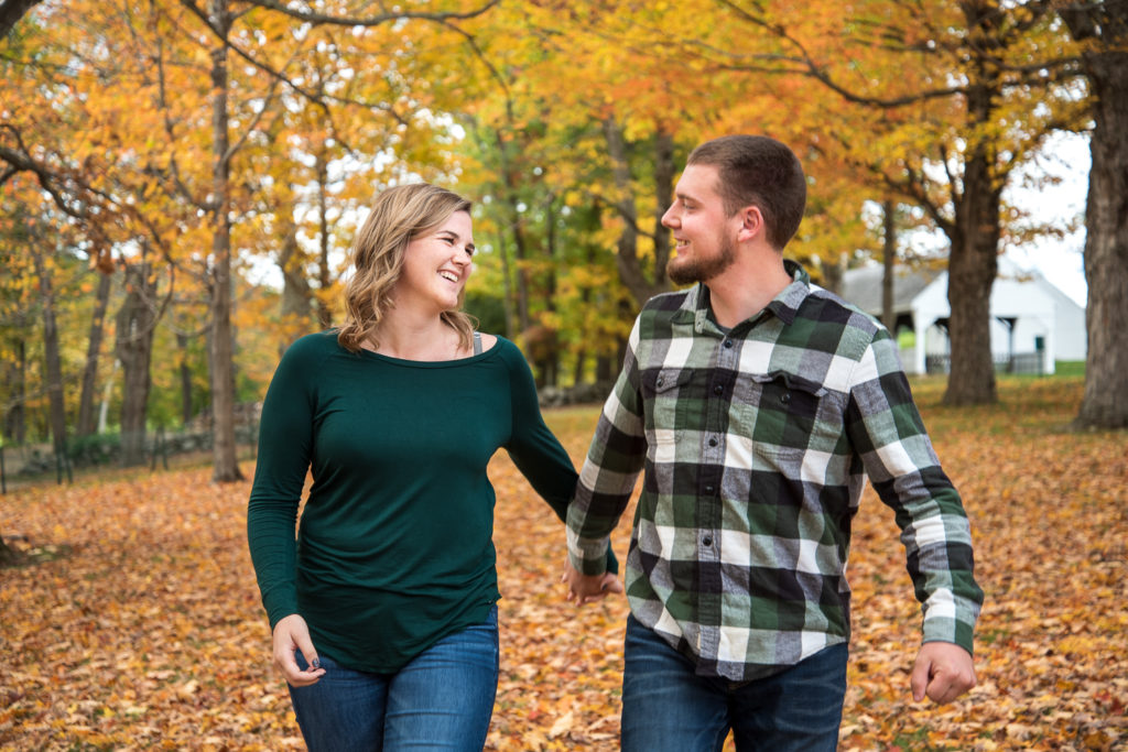 Best of 2021 Engagement couple walking hand in hand with fall foliage behind them in field 