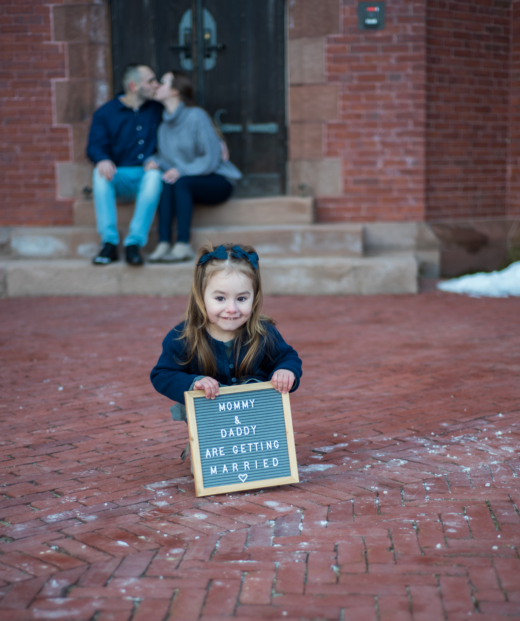 daughter holding sign saying mommy and daddy are getting married, while parents kiss in background