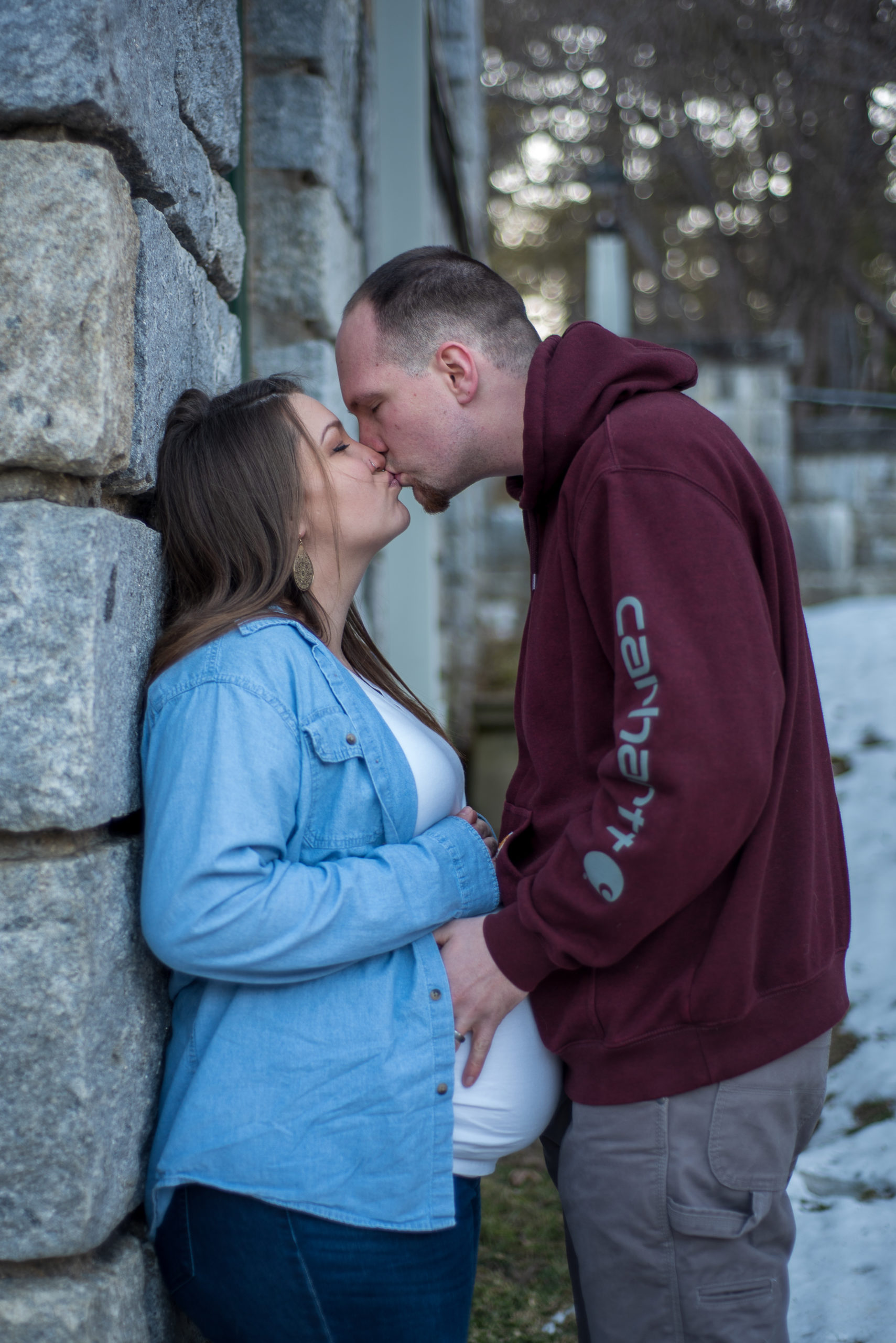 mom leaning against a wall, husband holding pregnant belly and kissing mom