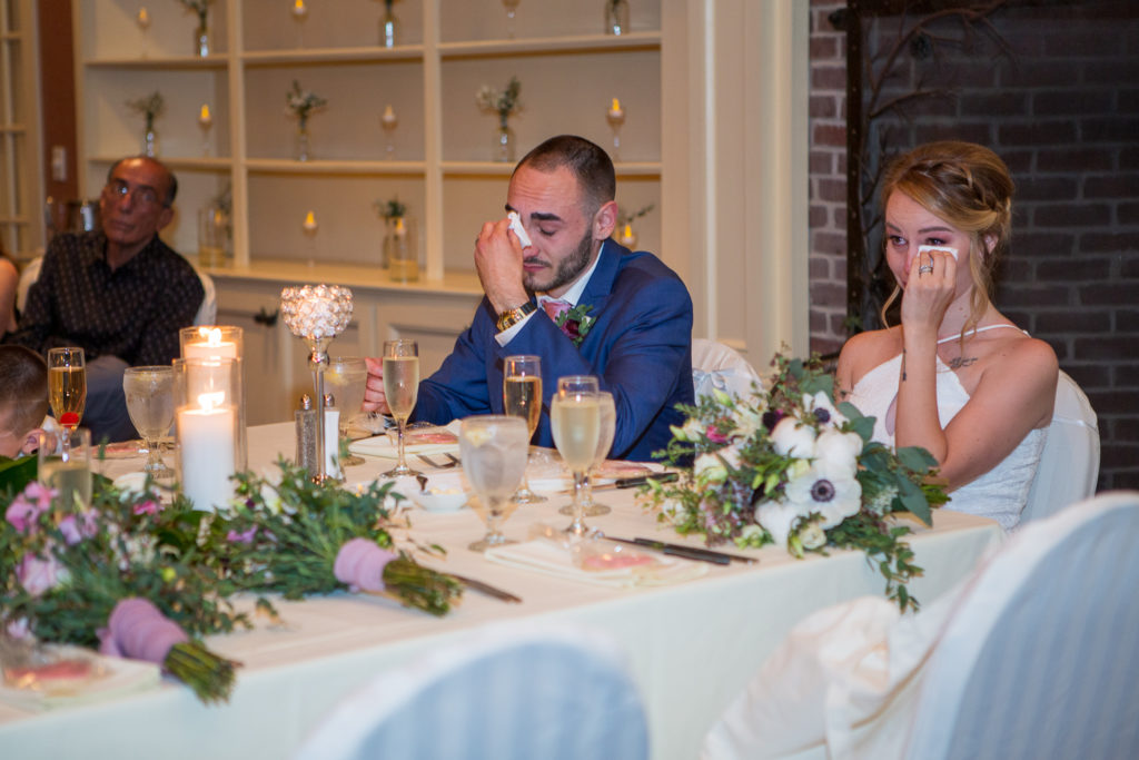 bride and groom wiping tears from their eyes as they listen to the bridal party give the wedding speeches 