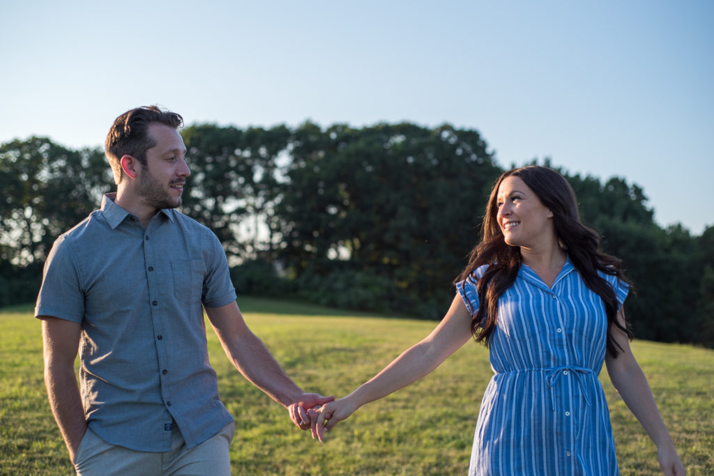 couple holding hands and smiling at each other as they walk in a field during sunset