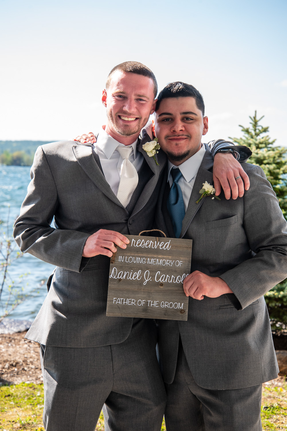 Groom and best man holding a sign in memory of the father of the groom after the lakeside wedding