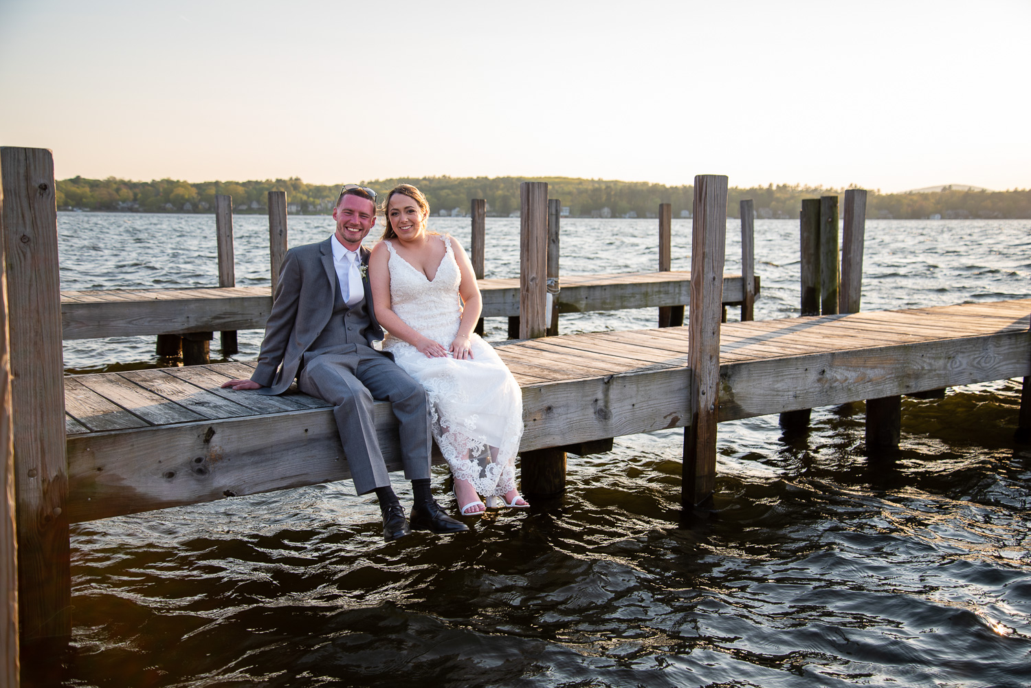 Bride and groom sitting on the edge of the dock with their feet hanging over the water at the lakeside wedding