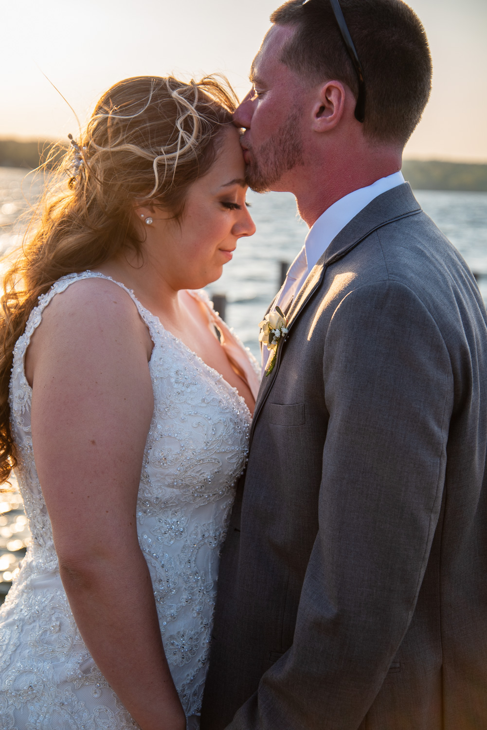 Groom kissing the brides forehead while standing on a dock with the sun setting at the lakeside wedding