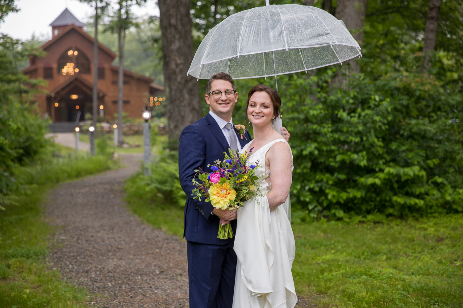 Bride and groom standing under umbrella in front of the barn at Old Saco Inn at their meadow wedding